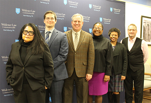 Six panelists stand together at the Dean's Diversity Forum at Widener Law Commonwealth, posing in front of a branded backdrop. From left to right: Guadalupe T. Luna, visiting professor of law, wears a black blazer and glasses; Ely Zeigler, a Center for Equity & Justice fellow, wears a blue suit and tie; Michael Hussey, associate professor of law, wears a brown plaid blazer and a light-colored shirt; Danielle M. Conway, dean at Penn State Dickinson Law, wears a purple dress and black blazer; Kellyn O. McGee, associate professor of law and faculty director for the Center for Equity & Justice, wears a black outfit with a pink top underneath; and Dean andre douglas pond cummings wears a black vest over a white dress shirt.