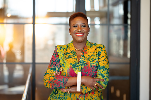 Image of Bunmi Emenanjo, a woman wearing a vibrant jacket and pants stands confidently in front of a large window, showcasing her stylish outfit.