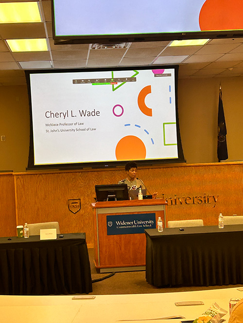 Cheryl L. Wade, McNeece Professor of Law at St. John’s University School of Law, speaks at a podium in a lecture hall at Widener University Commonwealth Law School. A large presentation slide with her name and title is displayed on the screen behind her. The lecture room is set up with tables and chairs, and the Widener University Commonwealth Law School logo is visible on the podium.