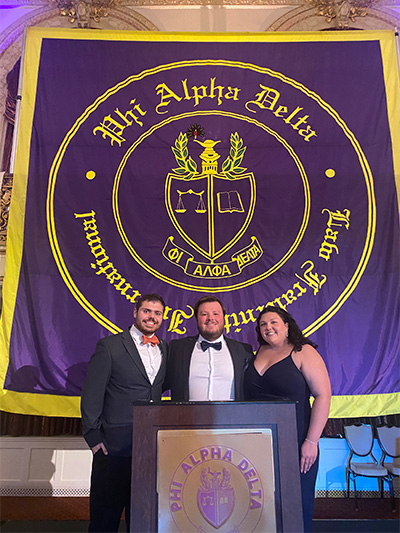 3 Phi Alpha Delta members standing in front of podium at conference in Pittsburgh with a purple Phi Alpha Delta flag in the background. 