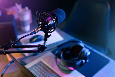 Image of podcast microphone and headphones alongside a laptop computer.