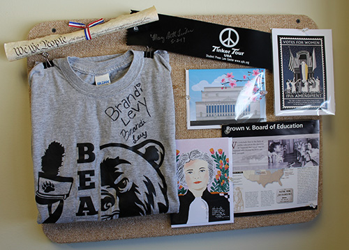 A corkboard display featuring a signed gray T-shirt with a bear mascot, a "We the People" parchment scroll, a "Tinker Tour" armband, a "Votes for Women" poster, a Brown v. Board of Education informational sheet, and illustrated postcards.