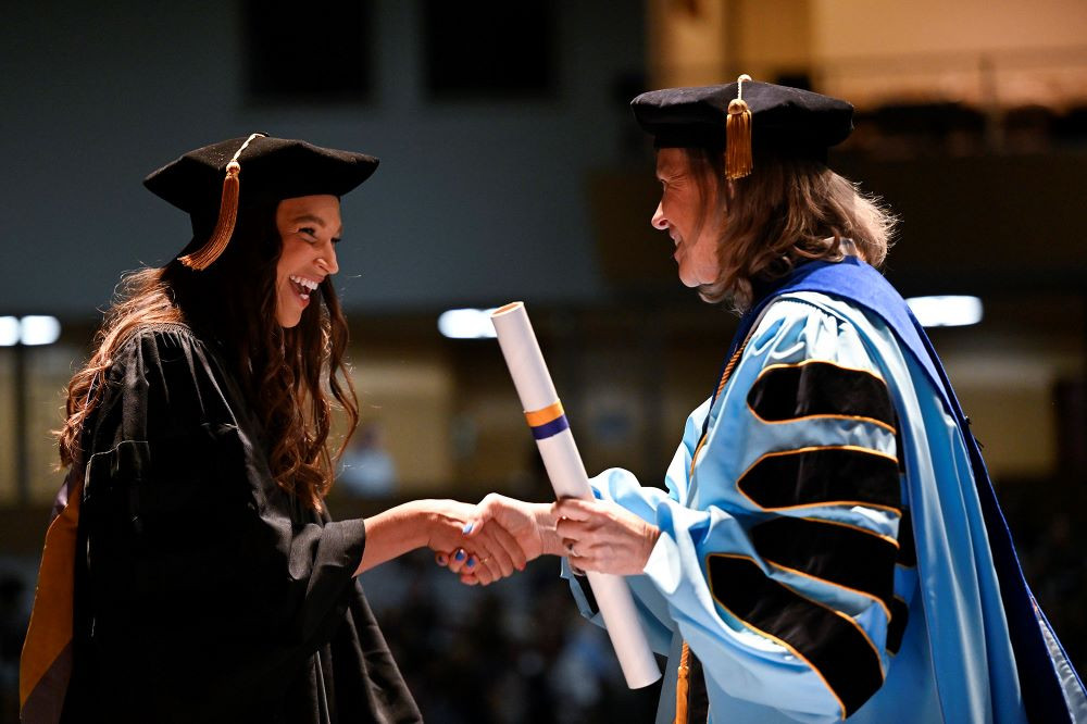 WLC graduate getting diploma from and shaking hands with President Robertson, both dressed in their regalia. 