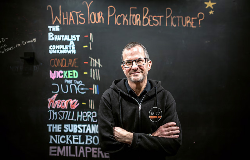 Todd Shill stands in front of a chalkboard tallying Best Picture picks.