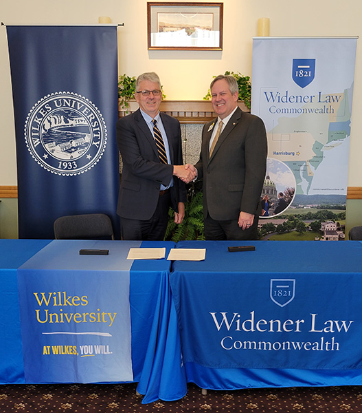Wilkes University representative and Dean Michael Hussey shaking hands near two blue tables after signing a 3+3 agreement