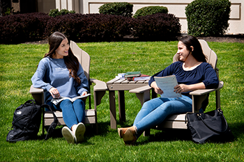 Image of two law students sitting on Adirondack chairs in front of the classroom building on campus on a bright sunny day. 