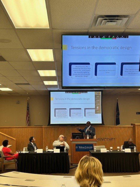 Professor Atiba Ellis presents at a podium in a Widener University Commonwealth Law School lecture hall. A slide titled 'Tensions in the Democratic Design' is displayed on two screens behind him, with text boxes detailing aspects of democratic challenge