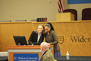 Natalie Solano and Kaylee Caprari standing in front of the Wolfberg Courtroom at the podium introducing Michael Consedine