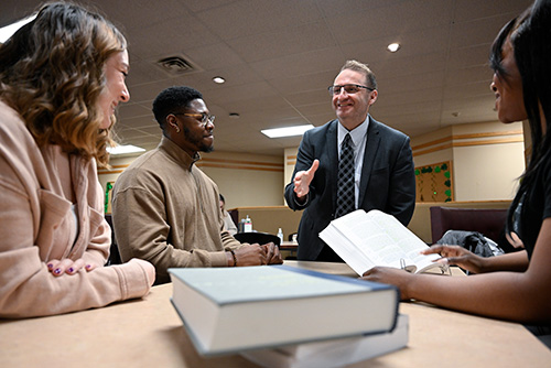 Dean andre cummings interacting with a group of students seated at a table, engaging in conversation with open books in the foreground, promoting learning and connection