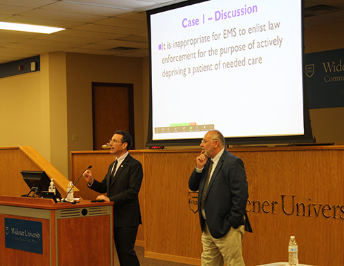 Two presenters at Widener Law Commonwealth lead a discussion during an EMS symposium. A presentation slide on the screen reads, 'It is inappropriate for EMS to enlist law enforcement for the purpose of actively depriving a patient of needed care.