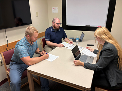 Three students sit in a mediation simulation around a table with laptops open. 