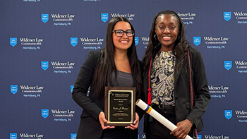Image of Emilie Baerga and Tomyra Wilson standing in front of the Widener Law Commonwealth branded step and repeat. They are smiling and holding their awards. 