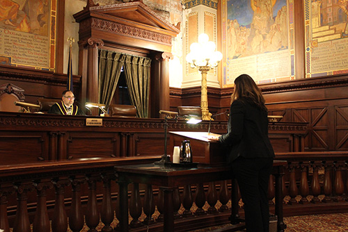 Alaina Kotash standing at a podium reading the graduates names to be sworn in while Justice Kevin Brobson sits behind the PA Supreme Court bench. 