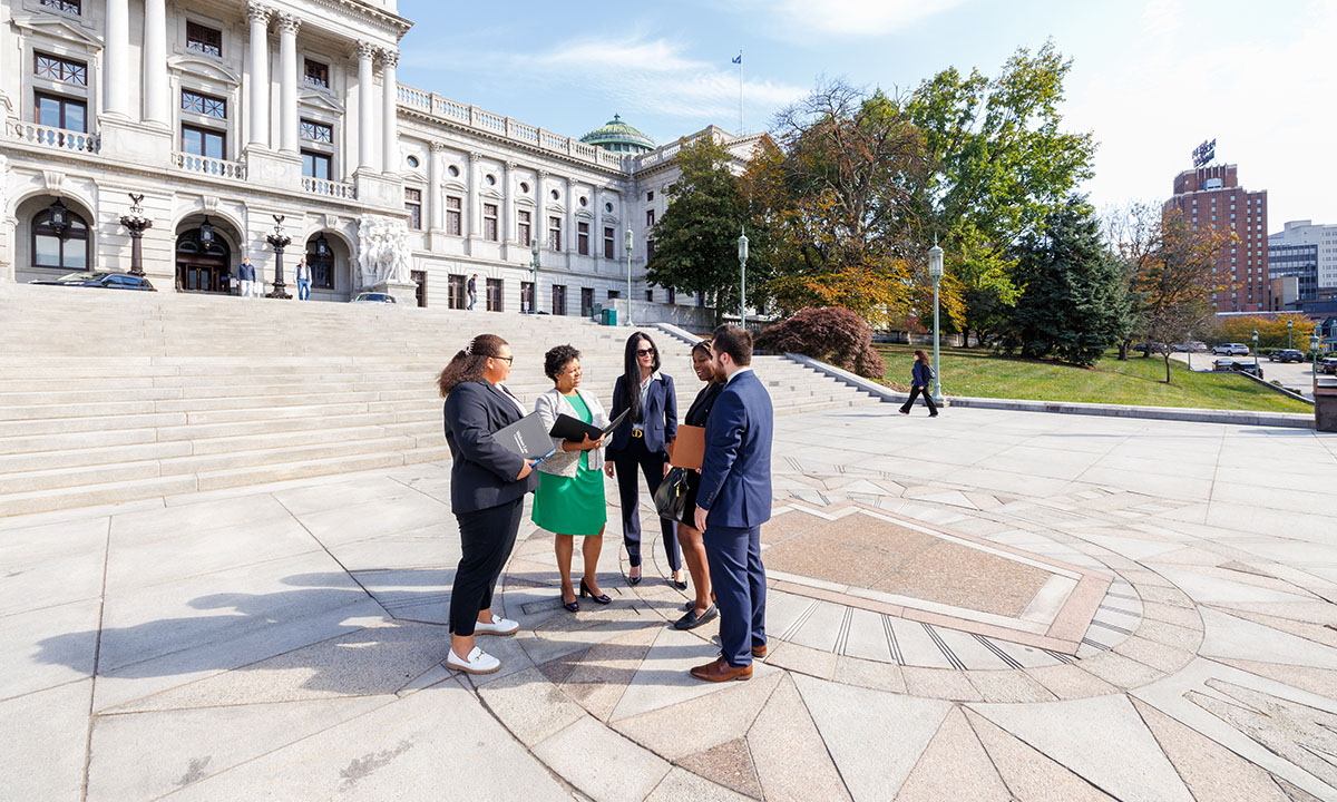 Kellyn McGee and three colleagues review documents together on the plaza in front of the Pennsylvania State Capitol, with the building's green dome visible behind them on a sunny fall day.