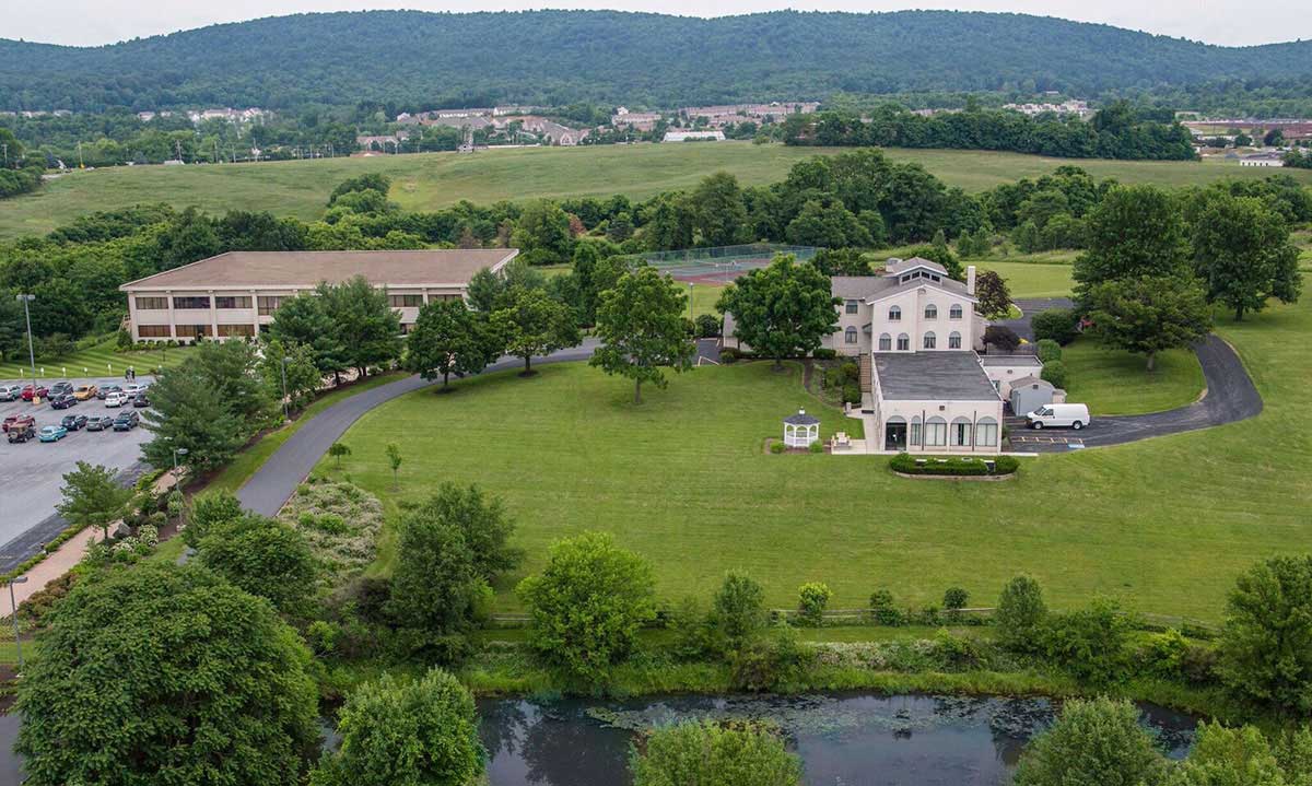 Aerial view of the Widener University Commonwealth Law School campus, showing the main building and adjacent structures set among expansive green lawns, mature trees, and a pond, with rolling hills and the broader Harrisburg region visible in the background.