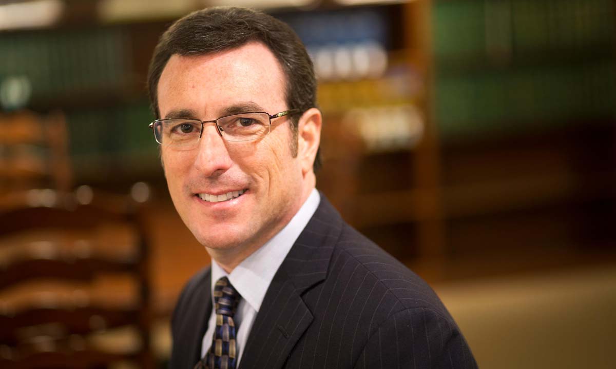 A smiling male attorney in a navy pinstripe suit and patterned tie, photographed in a law library setting.
