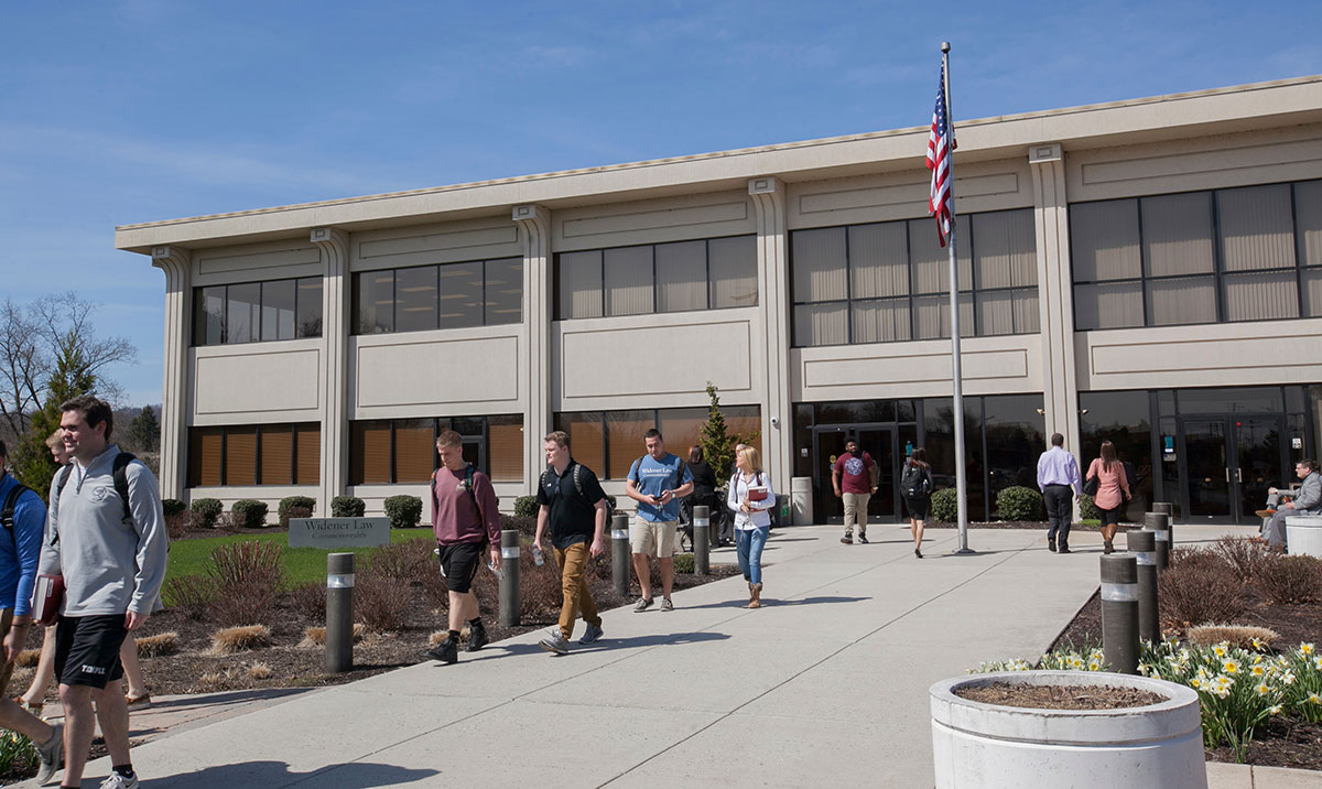 Students walk along the main entrance path of the Widener Law Commonwealth building on a sunny spring day, with an American flag flying out front and daffodils in bloom near the entrance.