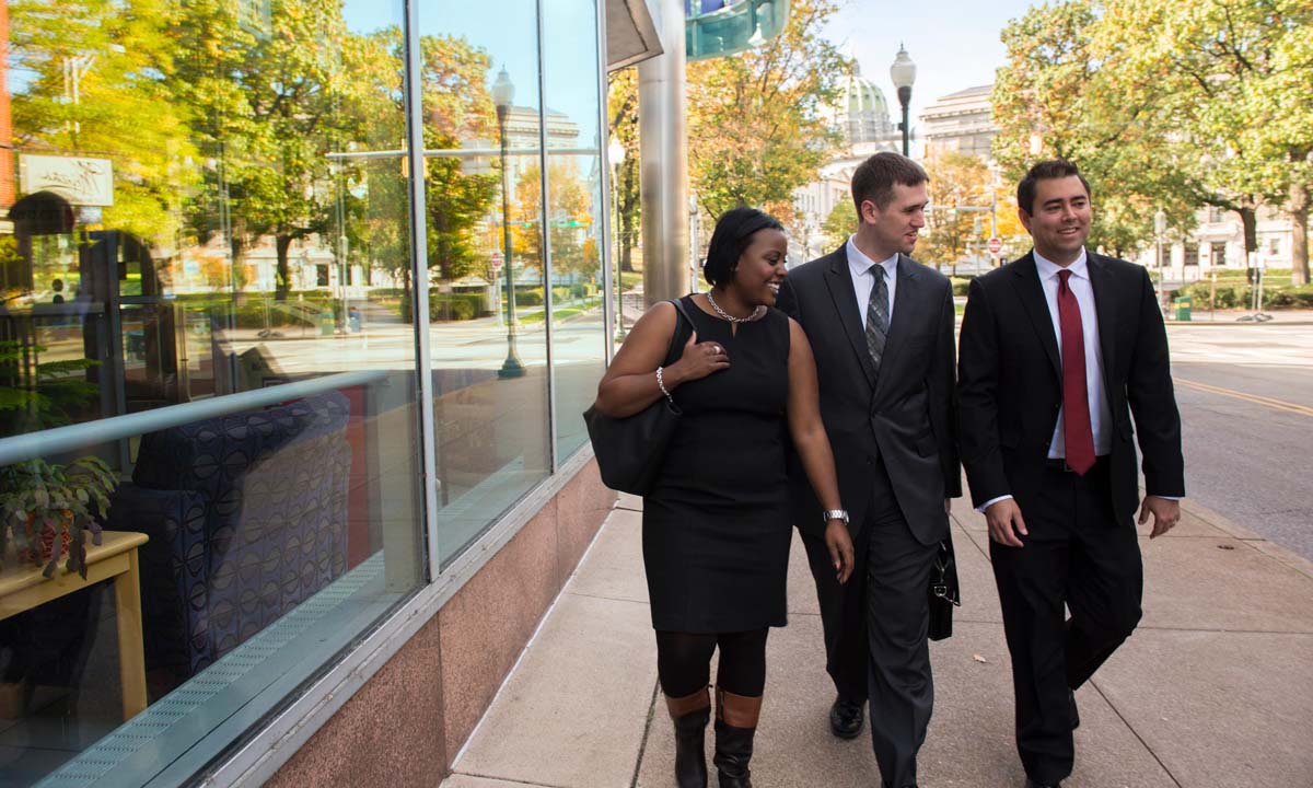 Three law students in professional attire walk and laugh together along a downtown Harrisburg sidewalk, with the Pennsylvania State Capitol dome visible among autumn trees in the background.