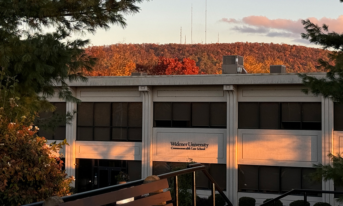 The Widener University Commonwealth Law School building glows in warm evening light against a backdrop of densely forested hills in full autumn color.