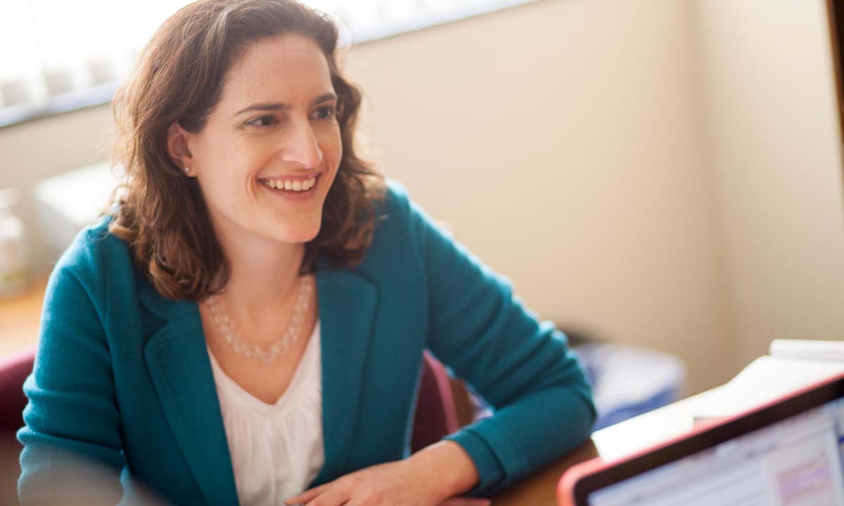 Jill Family smiles while seated at a desk in a bright office, wearing a teal blazer over a white top.