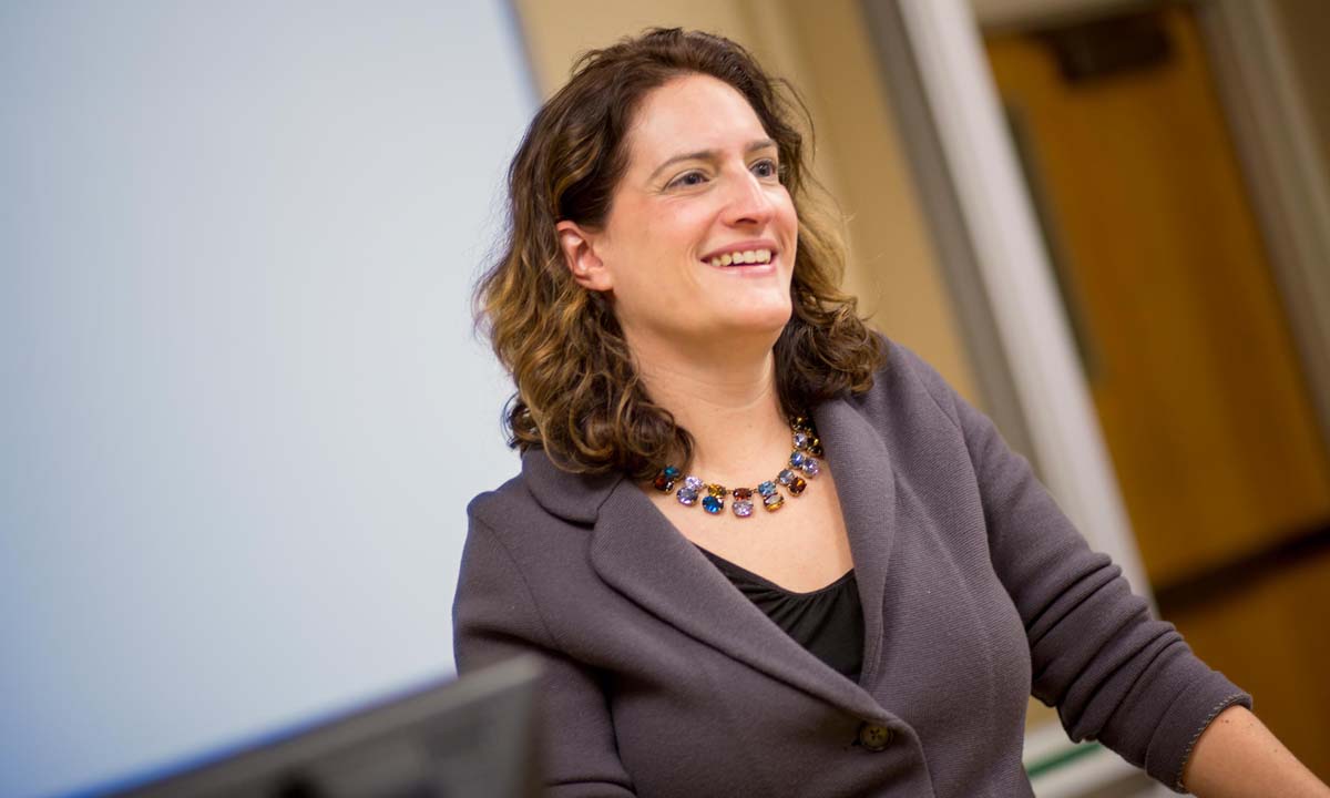 Jill Family smiles while teaching at the front of a classroom, wearing a gray blazer and colorful gemstone necklace.