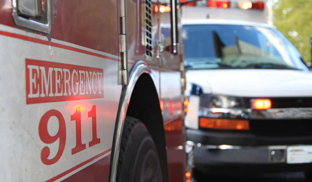 Close-up of an emergency vehicle marked "Emergency 911" with an ambulance visible in the background, lights activated.