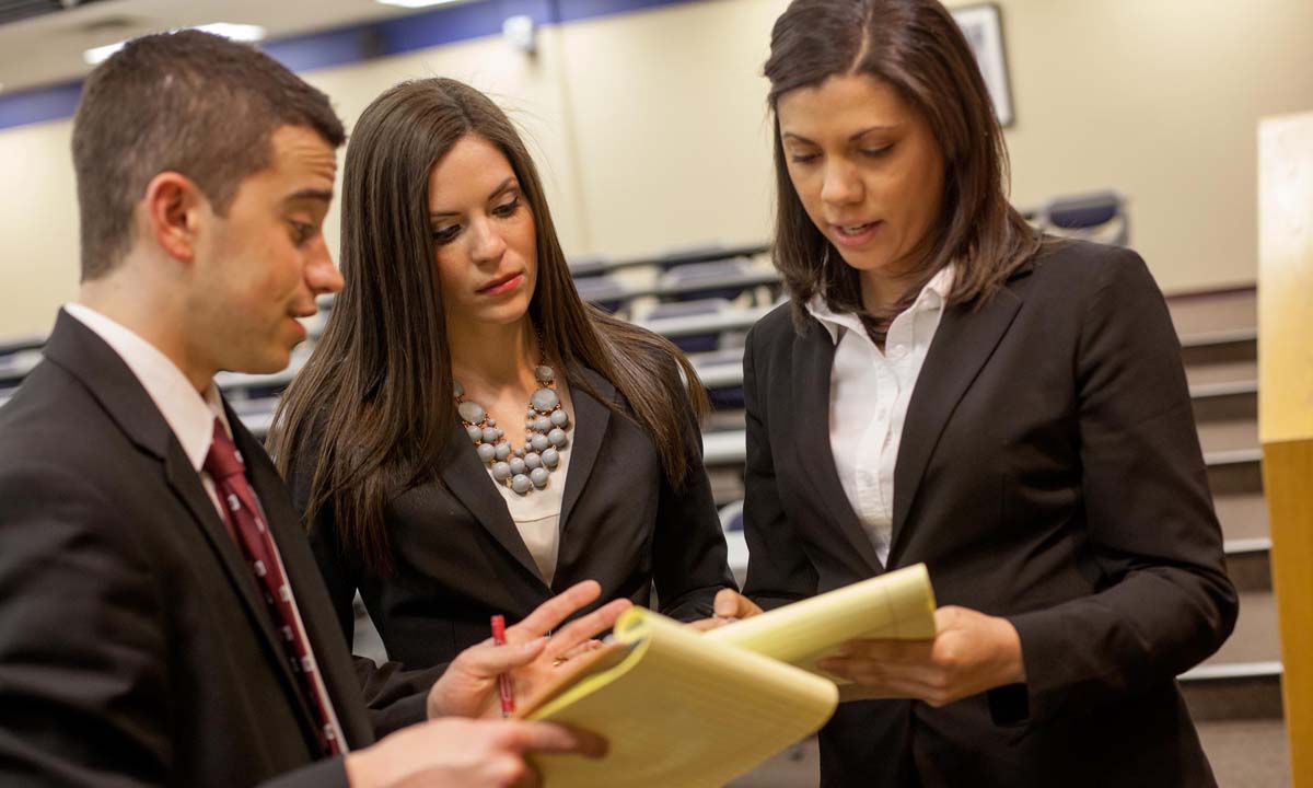 Three law students in professional business attire collaborate and review notes on yellow legal pads.