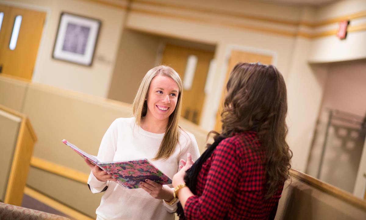 Two students stand in a university hallway talking while one student holds an open notebook.