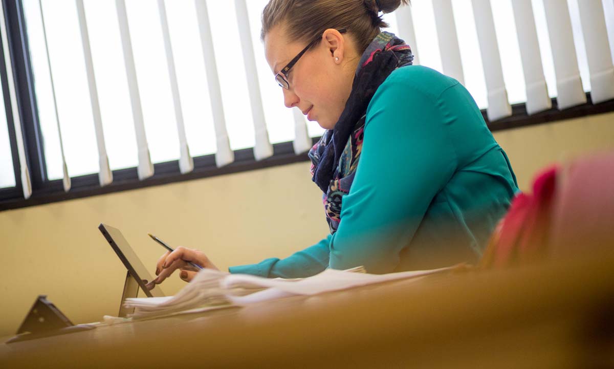 A student works intently on a tablet at a classroom desk, wearing a teal top and colorful scarf.