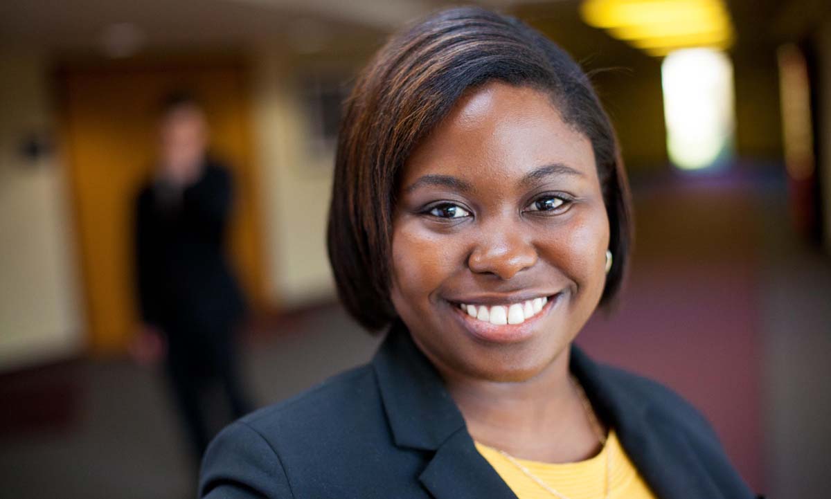 A smiling law student in a black blazer and yellow top, photographed in a campus hallway.
