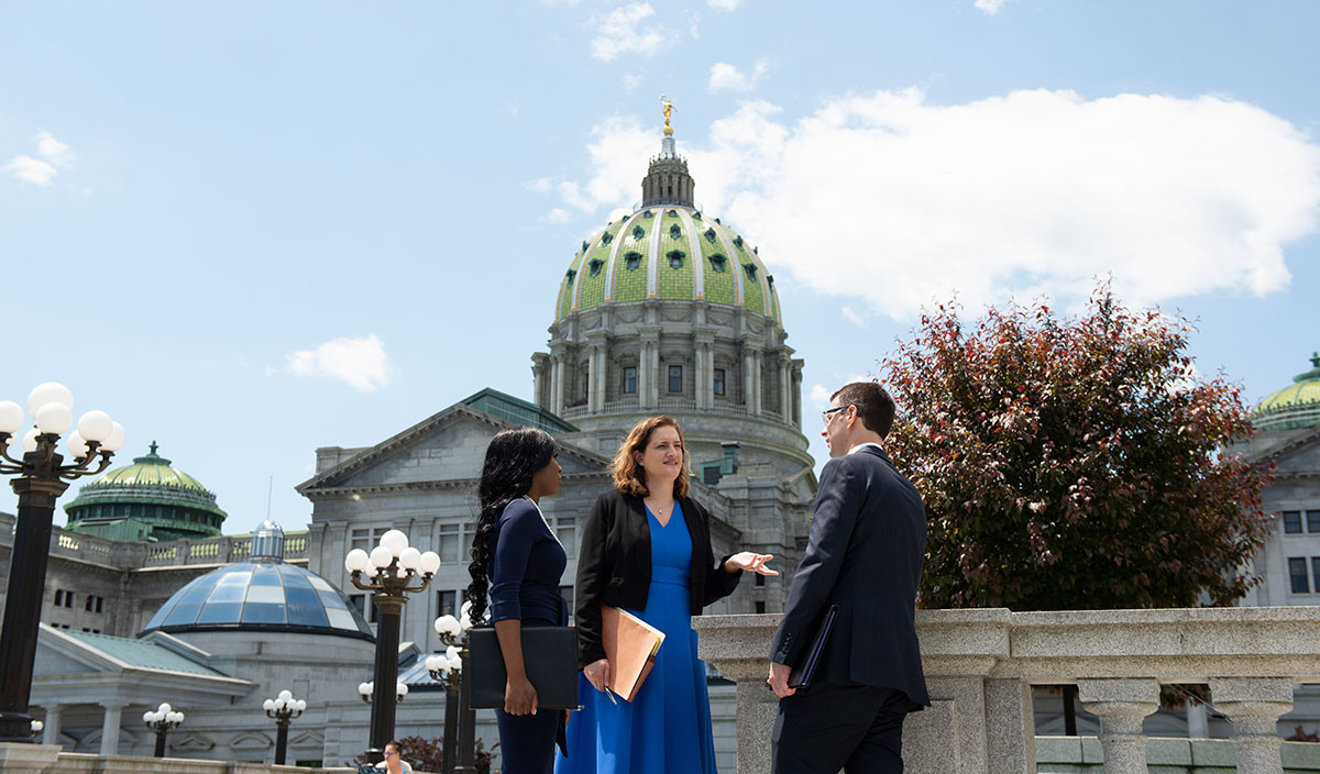 A faculty member converses with two students outside the Pennsylvania State Capitol building on a sunny summer day, with the Capitol's green dome rising prominently behind them.