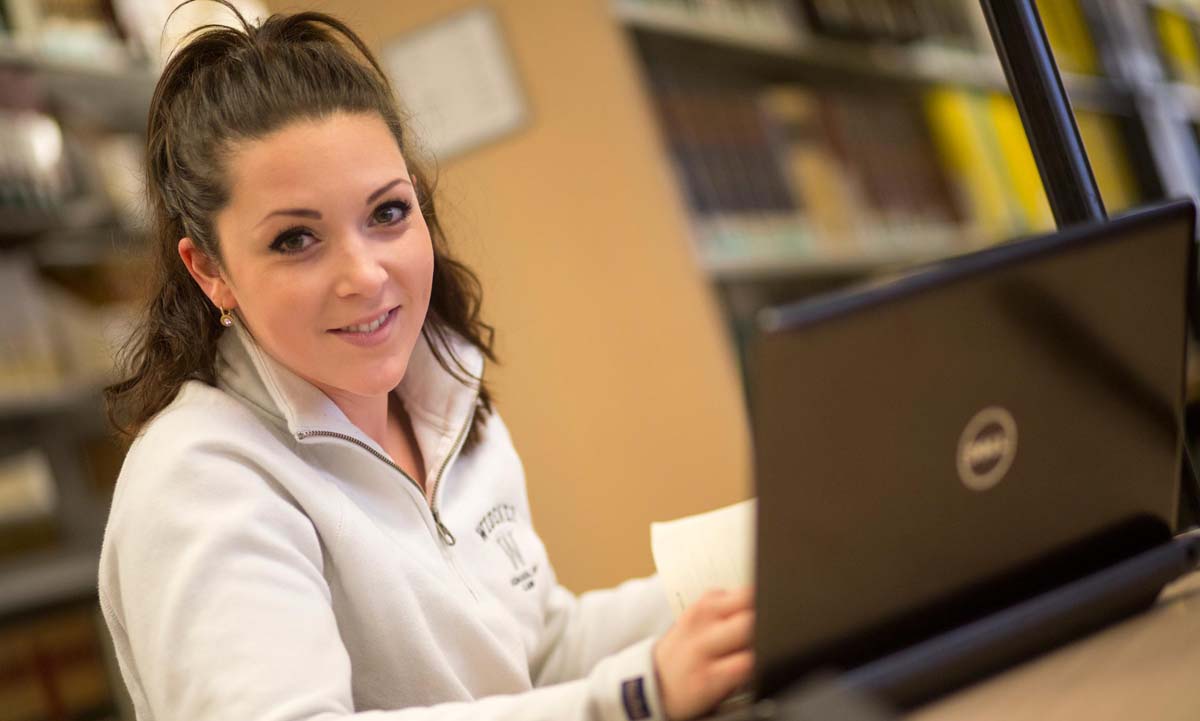 A law student in a Widener sweatshirt smiles while working on a laptop in the law library, holding a notecard with bookshelves visible in the background.