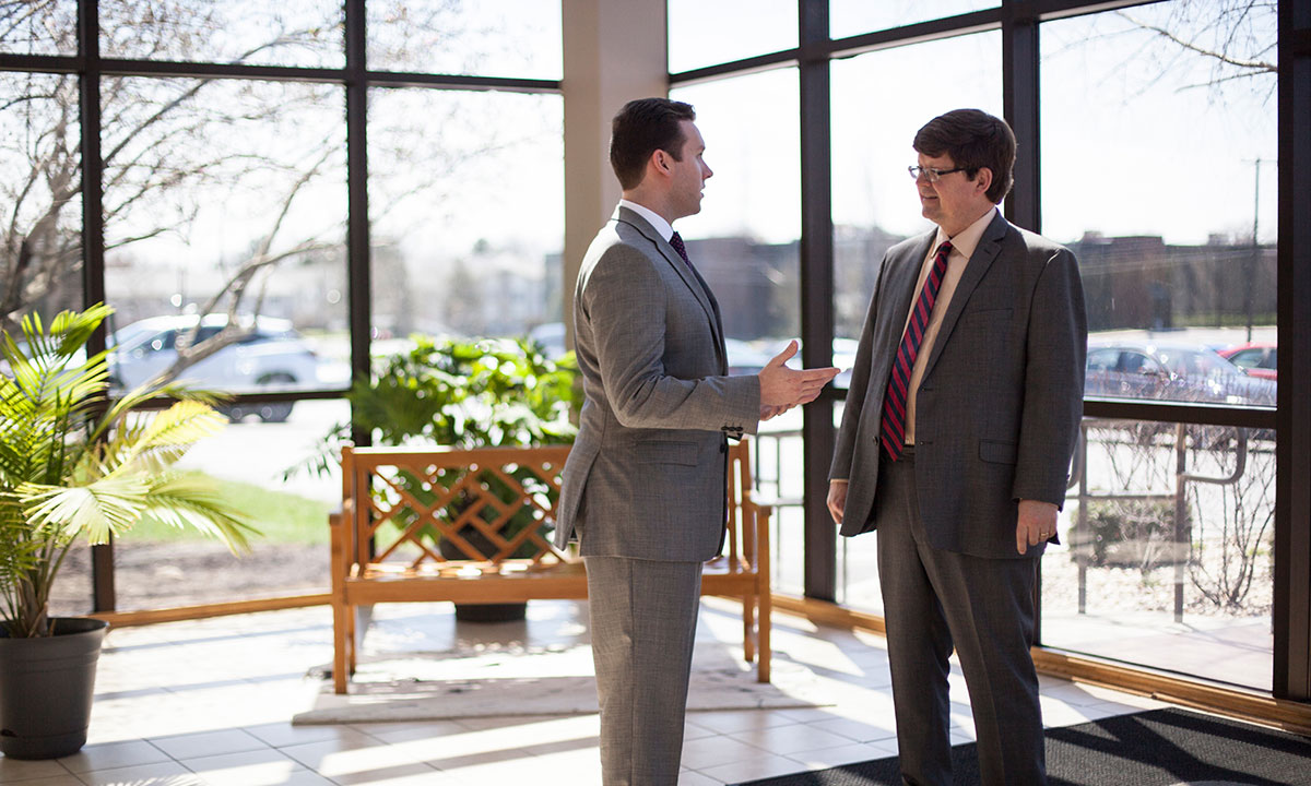 Two men in gray suits converse in a sunlit campus lobby, with large windows and bare winter trees visible in the background.