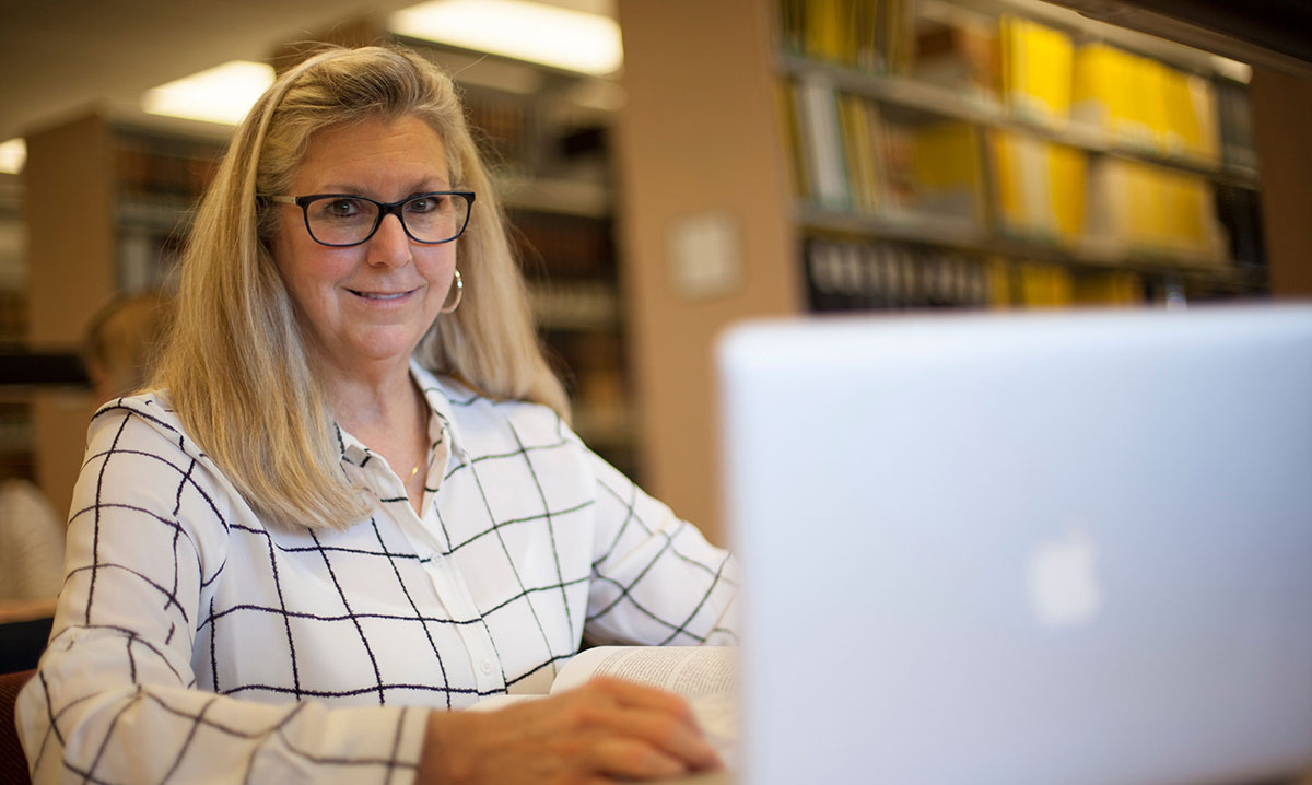A student works at a laptop in the campus law library, with shelves of legal volumes visible in the background.