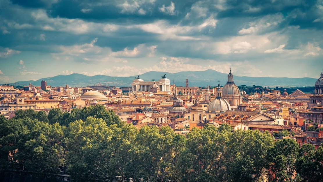 Panoramic view of Rome's historic skyline, with the Pantheon, the Altar of the Fatherland, and several church domes rising above the rooftops, with the Apennine mountains visible in the distance.