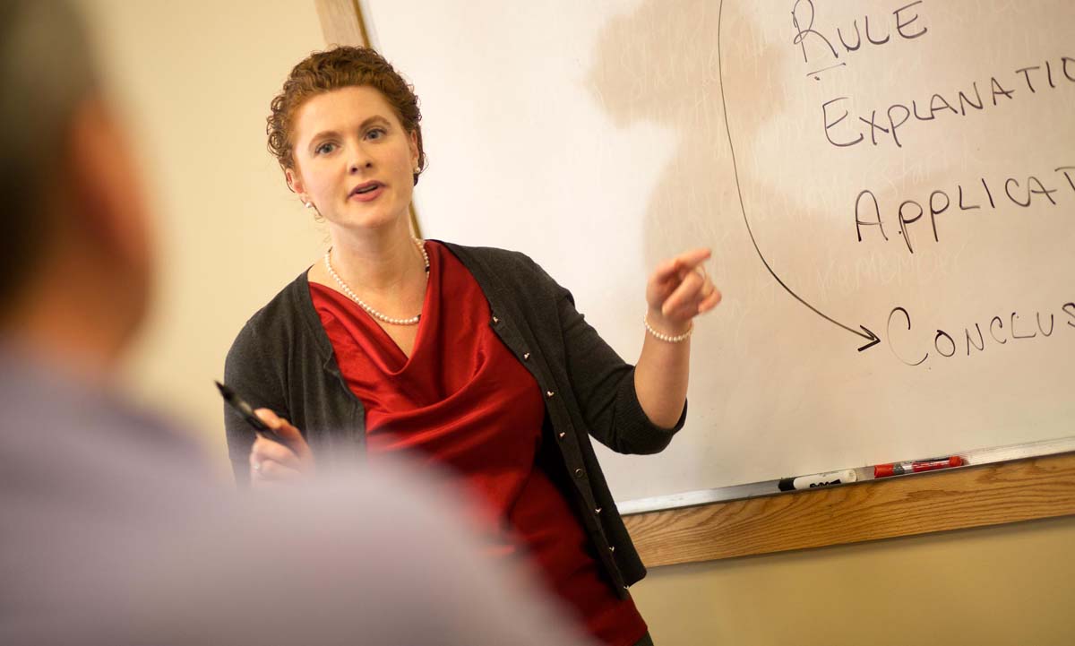 A faculty member gestures while teaching at a whiteboard with "Rule, Explanation, Application, Conclusion" written on it, wearing a red dress and black cardigan.