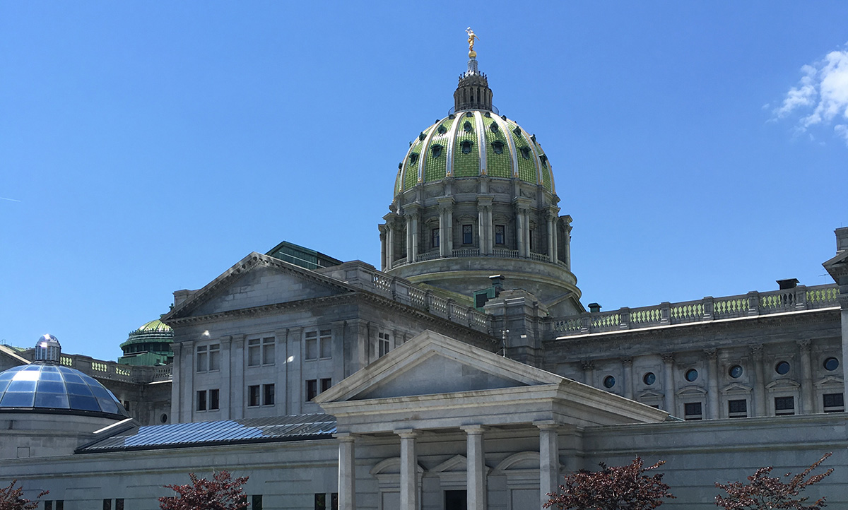 The Pennsylvania State Capitol building and its green dome rise against a clear blue sky, with a colonnaded entrance visible in the foreground.