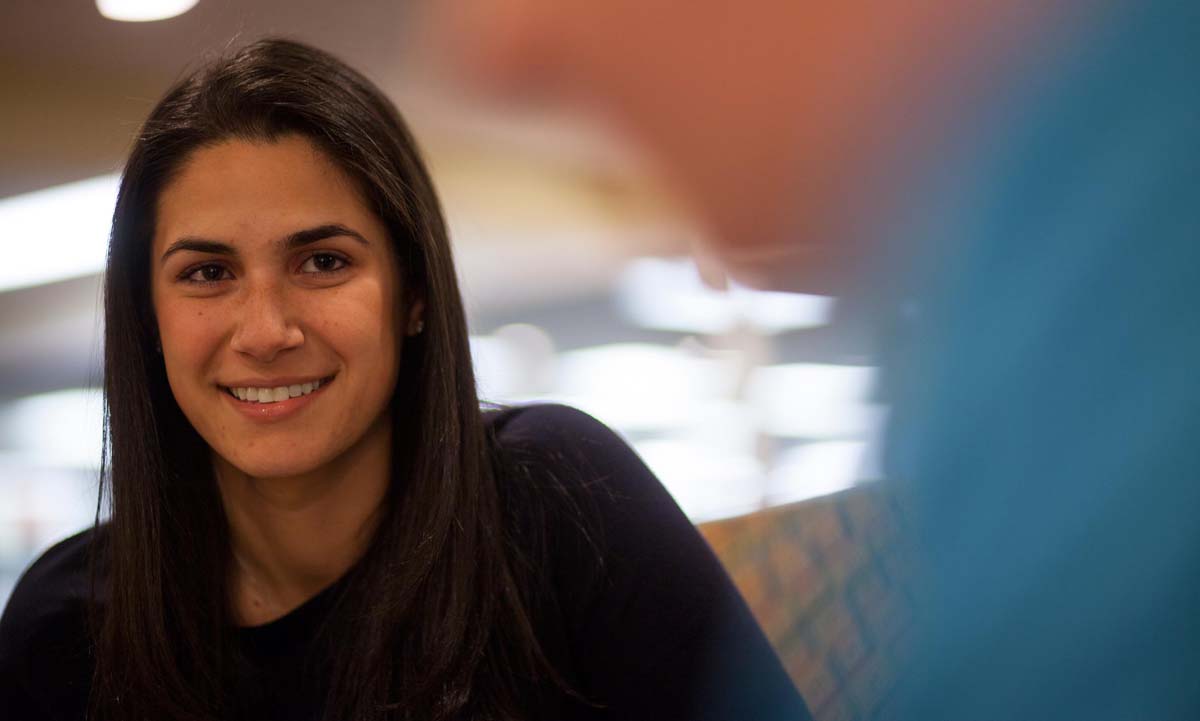 A student smiles in a close-up portrait in the campus law library, with the colorful lounge seating visible in the blurred background.