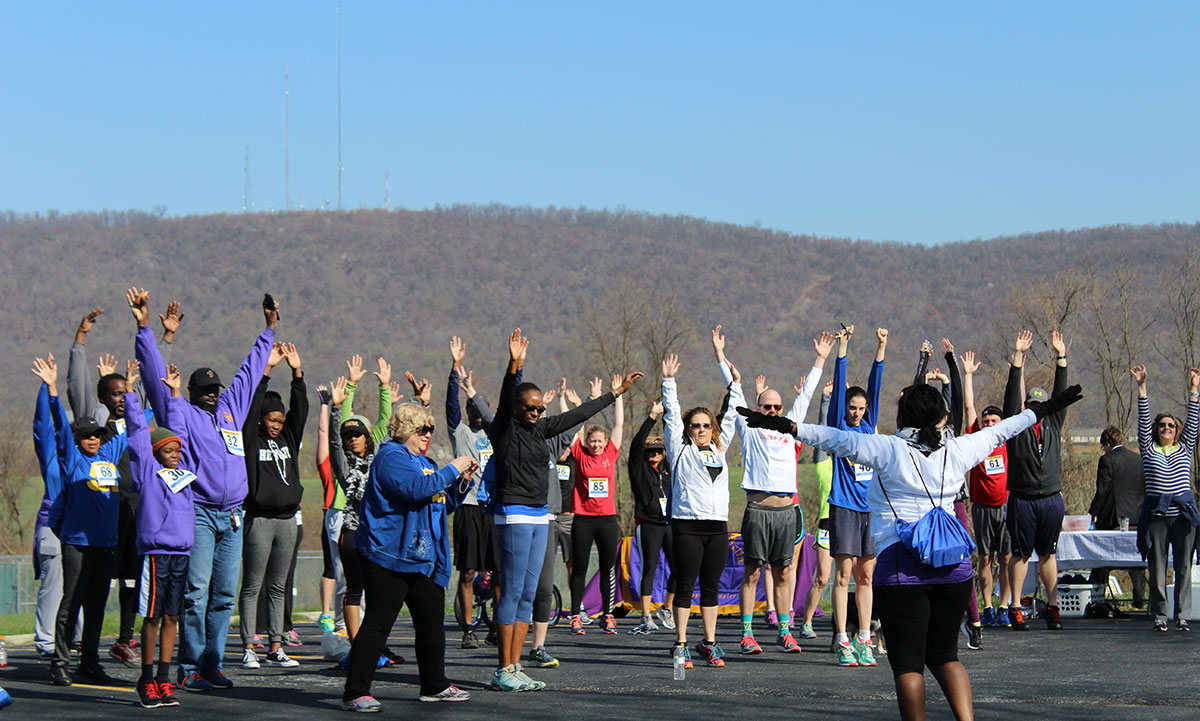 A large group of participants wearing race bibs stretch with arms raised during a pre-race warmup on a clear fall day, with tree-covered hills visible in the background.