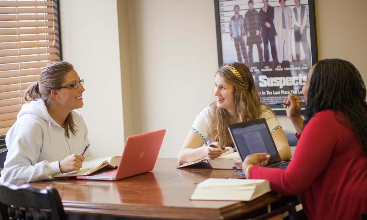 Three students sit around a table in a study room, discussing coursework while using notebooks and laptops.
