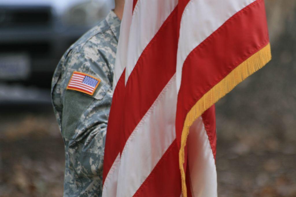 Close-up of a soldier in camouflage uniform holding an American flag with gold fringe, with a U.S. flag patch visible on the sleeve.