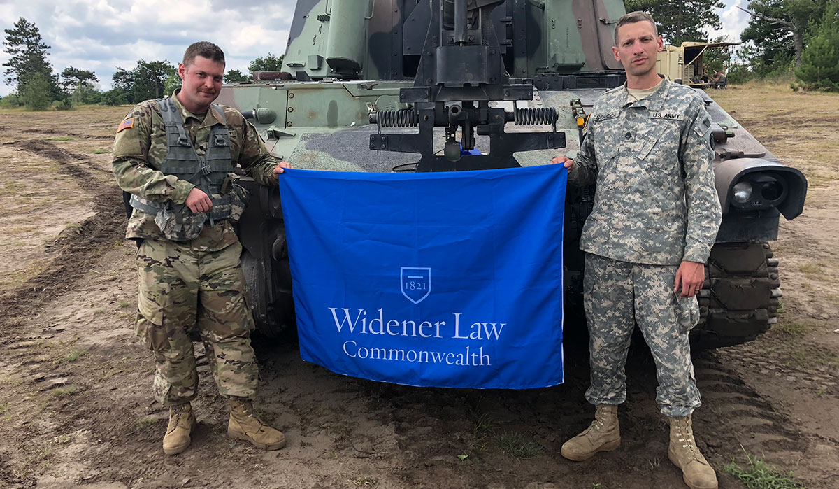 Two U.S. Army soldiers in uniform hold a Widener Law Commonwealth flag in front of a military vehicle in an outdoor field setting.