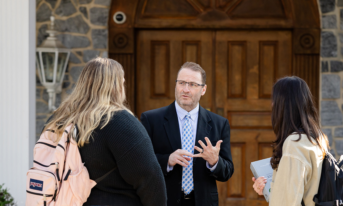 A faculty member gestures while speaking with two students outside the stone entrance of a campus building.