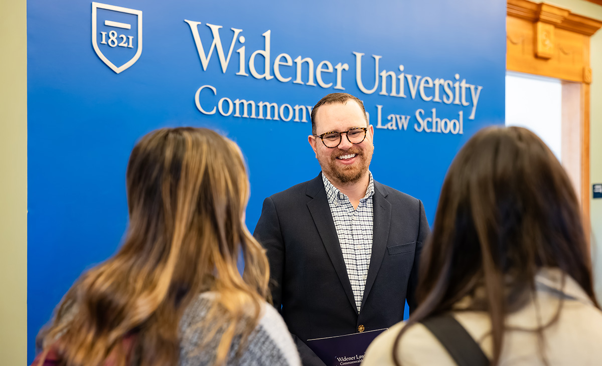 A smiling admissions staff member speaks with two prospective students in front of the Widener University Commonwealth Law School branded blue wall.