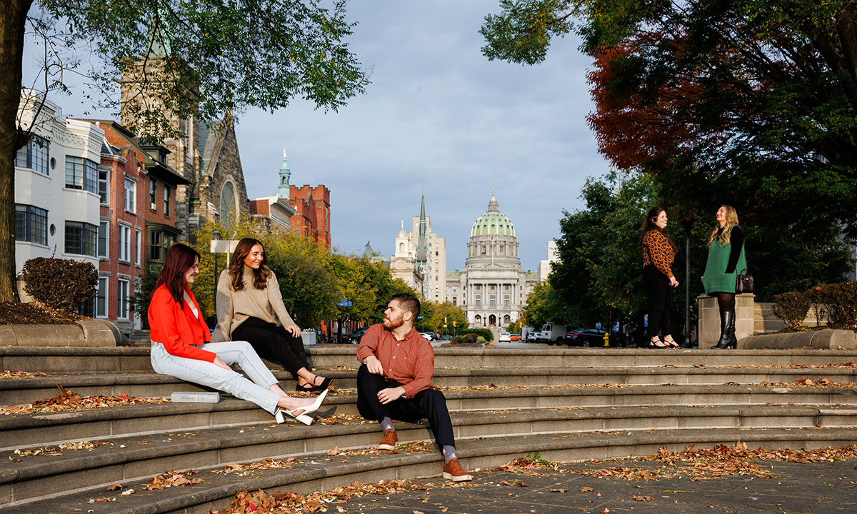 Law students relax on outdoor steps in downtown Harrisburg on an autumn day, with the Pennsylvania State Capitol dome and historic architecture visible along the tree-lined boulevard behind them.