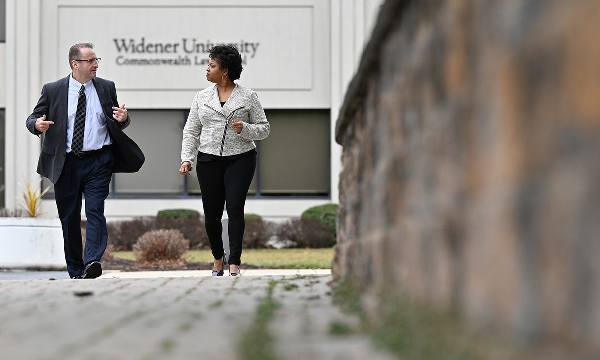 andré douglas pond cummings, wearing a suit and tie, walks and converses with a colleague outside the Widener University Commonwealth Law School building on a cool, overcast day.