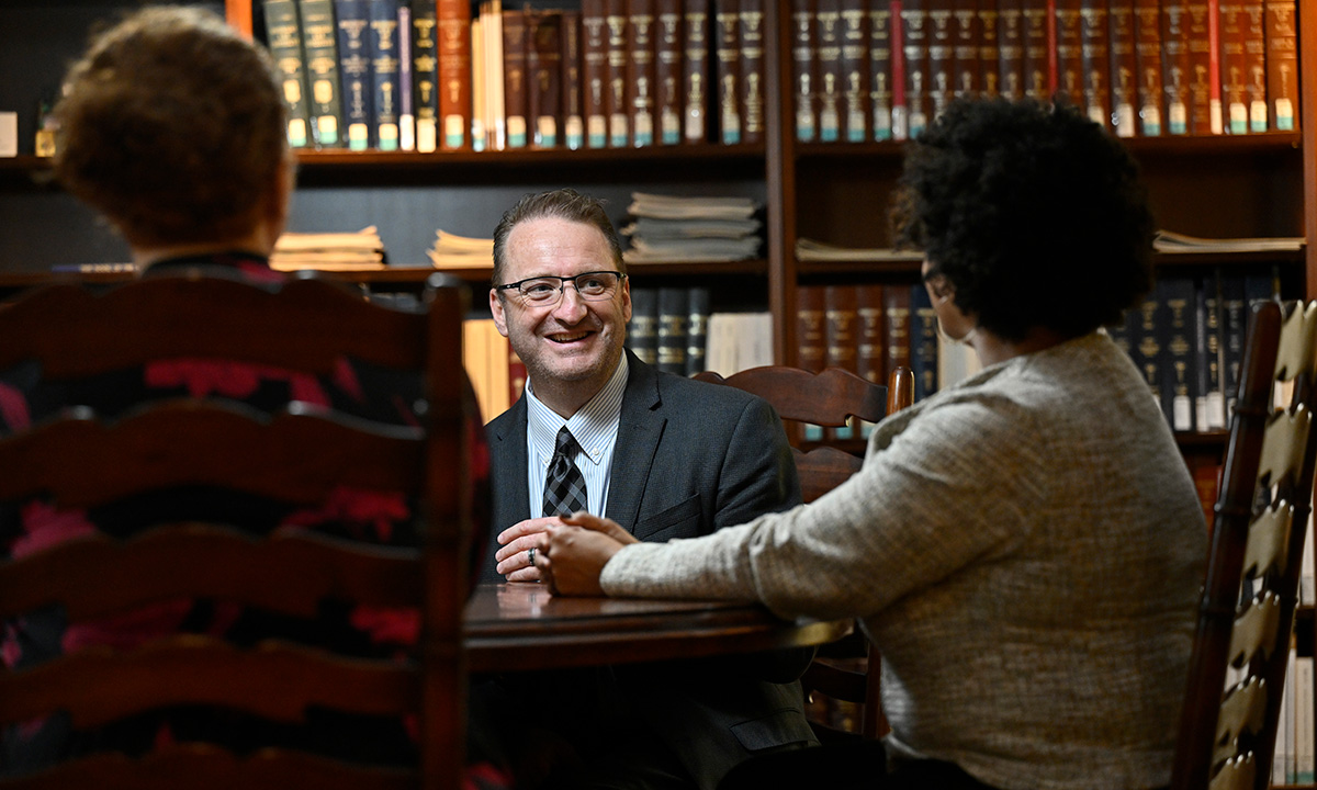 andré douglas pond cummings meets with two students at a table in a wood-paneled library room lined with law books, wearing a suit and tie.