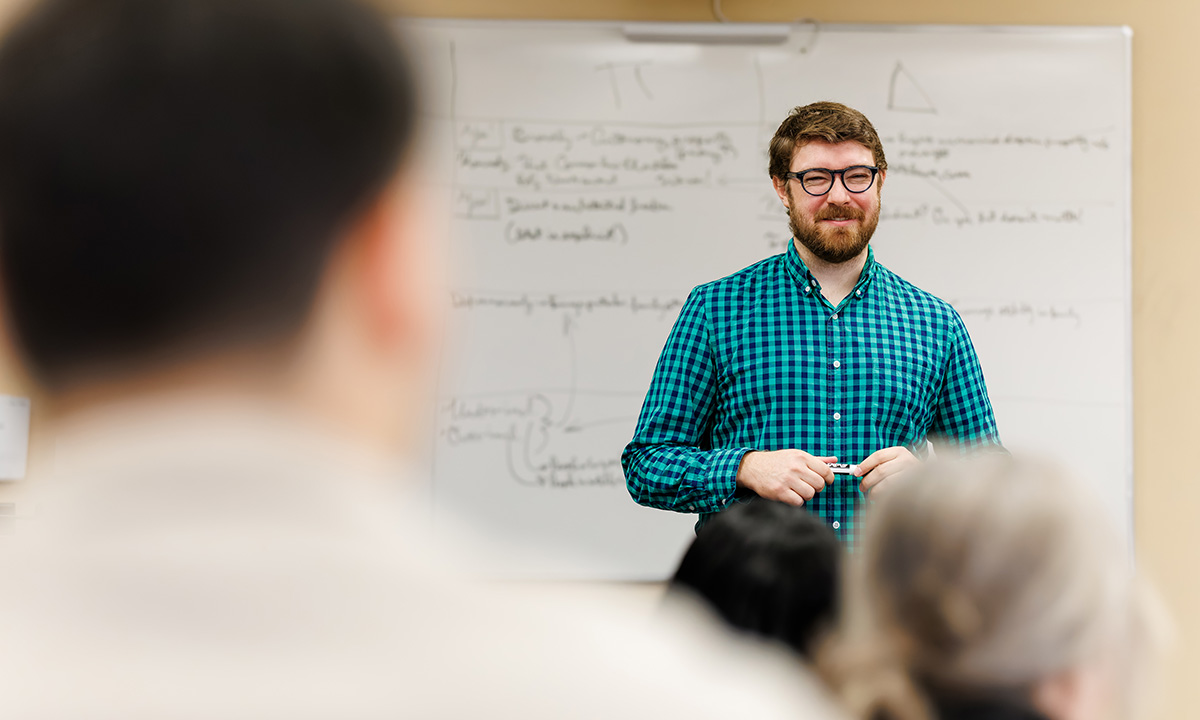 A faculty member stands at a whiteboard covered in notes, addressing students in a classroom, wearing a green plaid shirt and glasses.