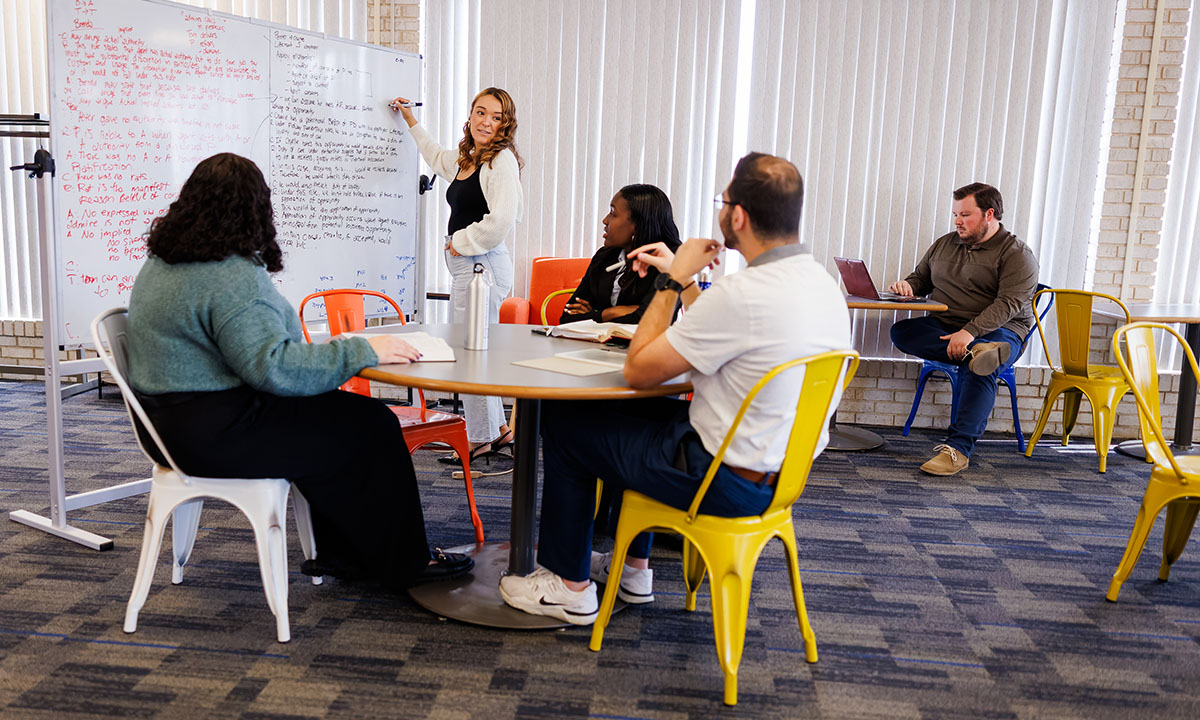 Law students collaborate in a study lounge, with one student leading the group at a whiteboard covered in notes while classmates follow along from colorful chairs around a round table.