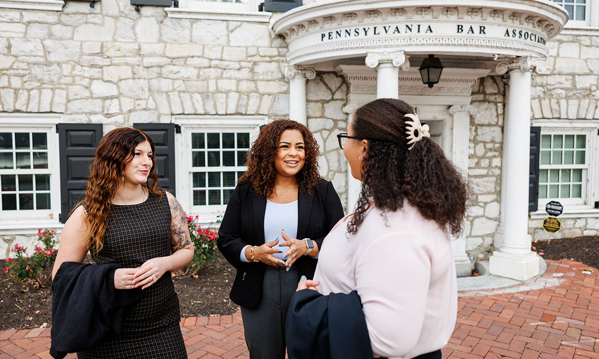 Three women converse outside the Pennsylvania Bar Association building, standing on a brick walkway in front of the organization's columned stone entrance.